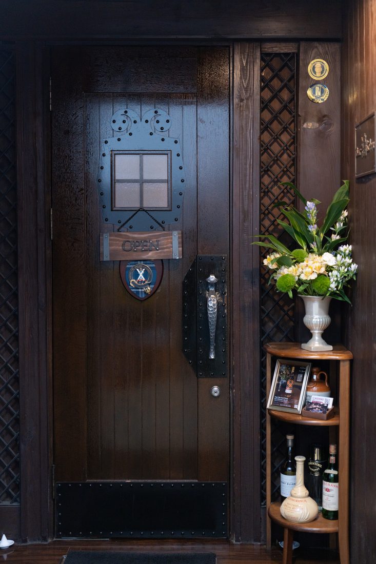 店舗入口の扉全景。小窓と真鍮ノブ、脇の棚にボトルと生花が並ぶ