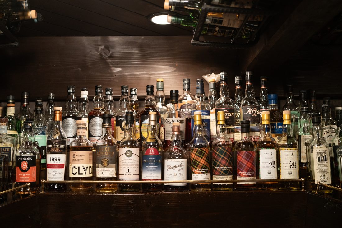 バックバーの棚に約20本のボトルが一列に並ぶ集合カット。中央にBar Kohnoオリジナルボトル「鴻」とGlenfarclas 2010 25年、Springbank 10やThe Maltman 1999、Kilchoman、Bowmoreなどが真横から整列している